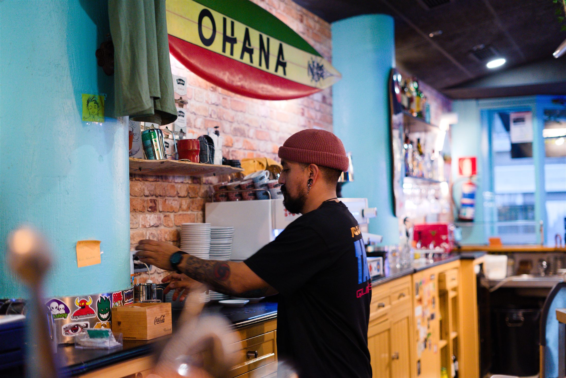 Un ohanero con gorro granate trabajando tras la barra, bajo la tabla de surf rasta con el nombre OHANA